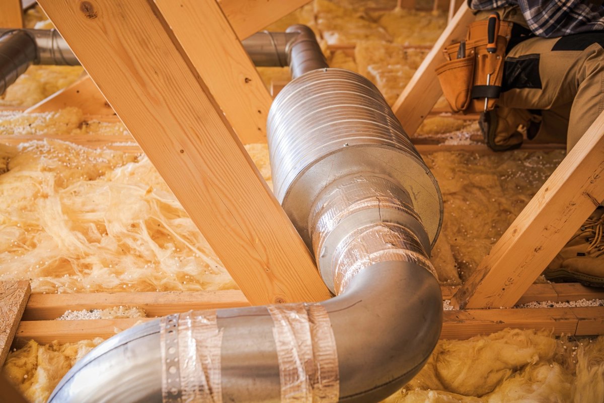 Metal air ducts run through an unfinished attic among wooden joists and yellow insulation.